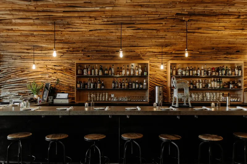 The bar at Bida Manda in Raleigh, with its signature stacked-driftwood wall, back-bar liquor shelves, Edison-bulb pendants, and a row of wooden bar stools