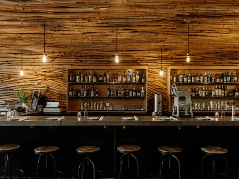 The bar at Bida Manda in Raleigh, with its signature stacked-driftwood wall, back-bar liquor shelves, Edison-bulb pendants, and a row of wooden bar stools