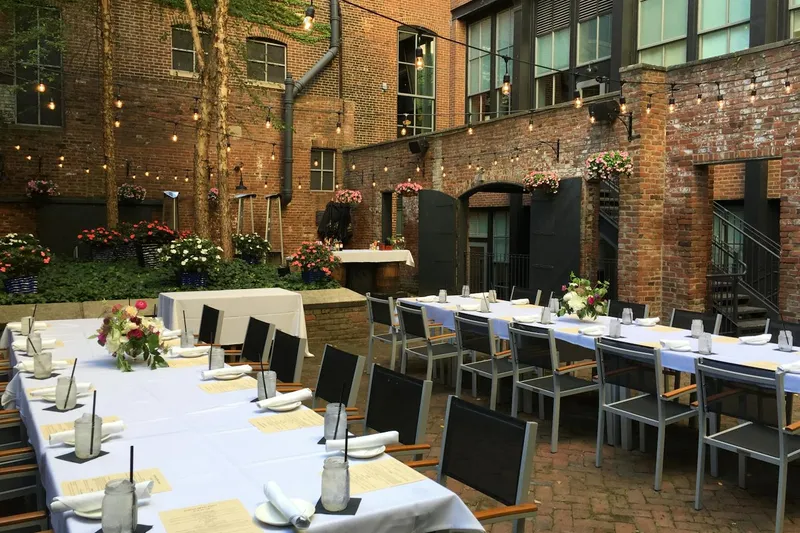 Outdoor brick courtyard patio at Mussel & Burger Bar Downtown in Louisville, set with long white-linen tables, black chairs, mason-jar water glasses and flowers under strung café lights