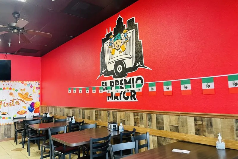 Dining room at El Premio Mayor in Fresno, with a hand-painted El Premio Mayor taco-truck mural on a red wall, wood-paneled wainscoting, wooden tables and chairs, and Mexican flag bunting