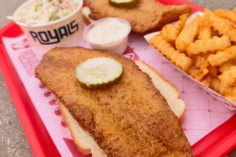 Red cafeteria tray at Royals Hot Chicken holding a fried fish fillet on white bread topped with a pickle, a branded cup of creamy coleslaw, a tub of tartar sauce and a basket of seasoned crinkle-cut fries