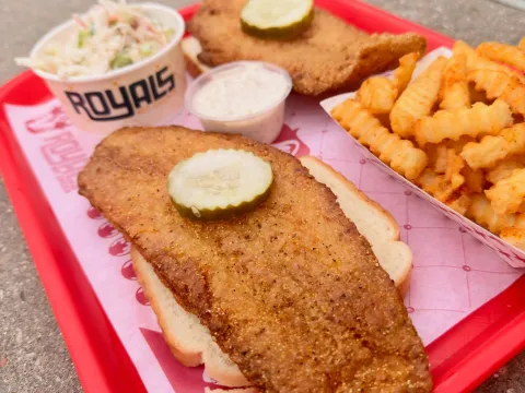 Red cafeteria tray at Royals Hot Chicken holding a fried fish fillet on white bread topped with a pickle, a branded cup of creamy coleslaw, a tub of tartar sauce and a basket of seasoned crinkle-cut fries