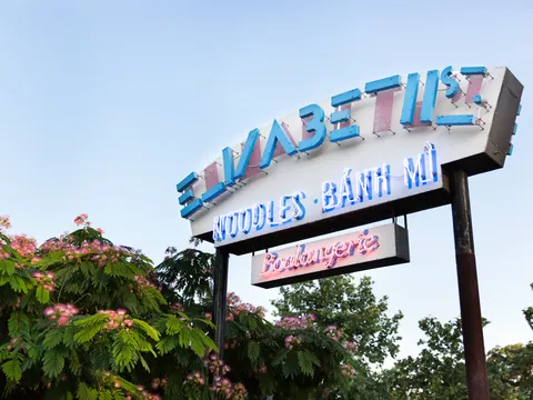 Elizabeth Street Café's neon street sign reading 'Noodles · Bánh Mì · Boulangerie' above mimosa trees in Austin