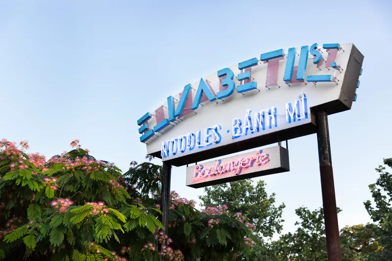 Elizabeth Street Café's neon street sign reading 'Noodles · Bánh Mì · Boulangerie' above mimosa trees in Austin
