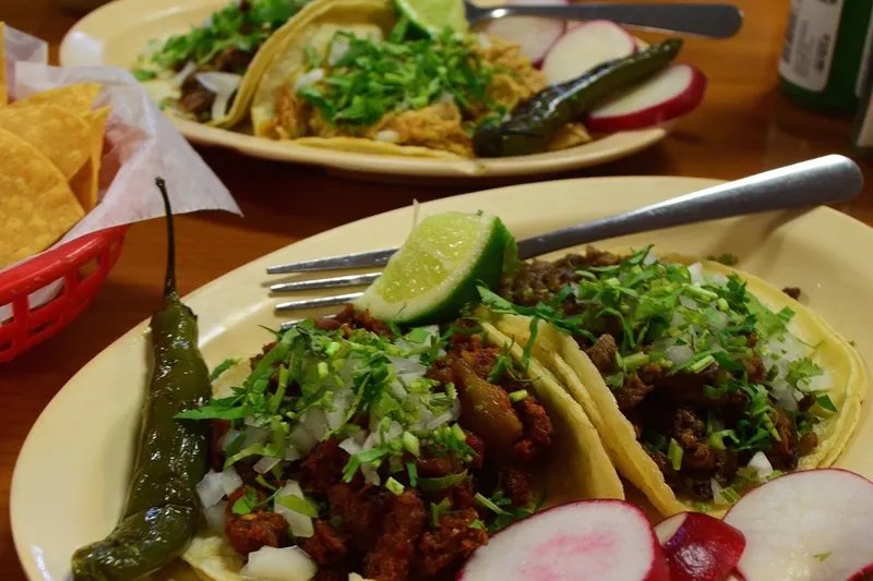 Two plates of street tacos at Taqueria La Guadalupana topped with chopped onion and cilantro, served with grilled jalapeños, radishes, lime and a basket of tortilla chips