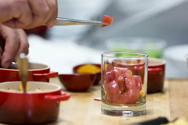 A cook at Seviche plating cubed raw tuna into a glass over citrus broth with tweezers, surrounded by small red cocotte dishes on a wooden cutting board in the kitchen