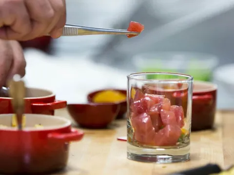 A cook at Seviche plating cubed raw tuna into a glass over citrus broth with tweezers, surrounded by small red cocotte dishes on a wooden cutting board in the kitchen