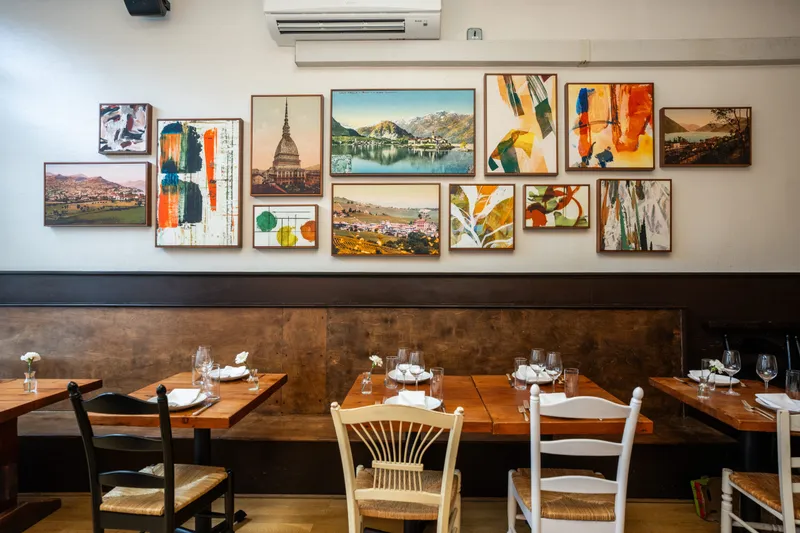 Dining room at Spinasse in Seattle, with set wooden tables and a bench banquette below a wall of framed Piedmont landscape and abstract art