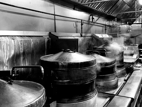 Black-and-white photo of the Jasmine Seafood kitchen line, with rows of large stacked bamboo-and-metal dim sum steamers venting steam