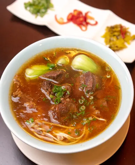 Bowl of Facing East Taiwanese beef noodle soup with braised beef, baby bok choy and scallions, served with side condiments