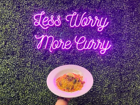 Plate of biryani held up in front of the pink "Less Worry More Curry" neon sign on a greenery wall at The Curry Modern Indian in Huntsville